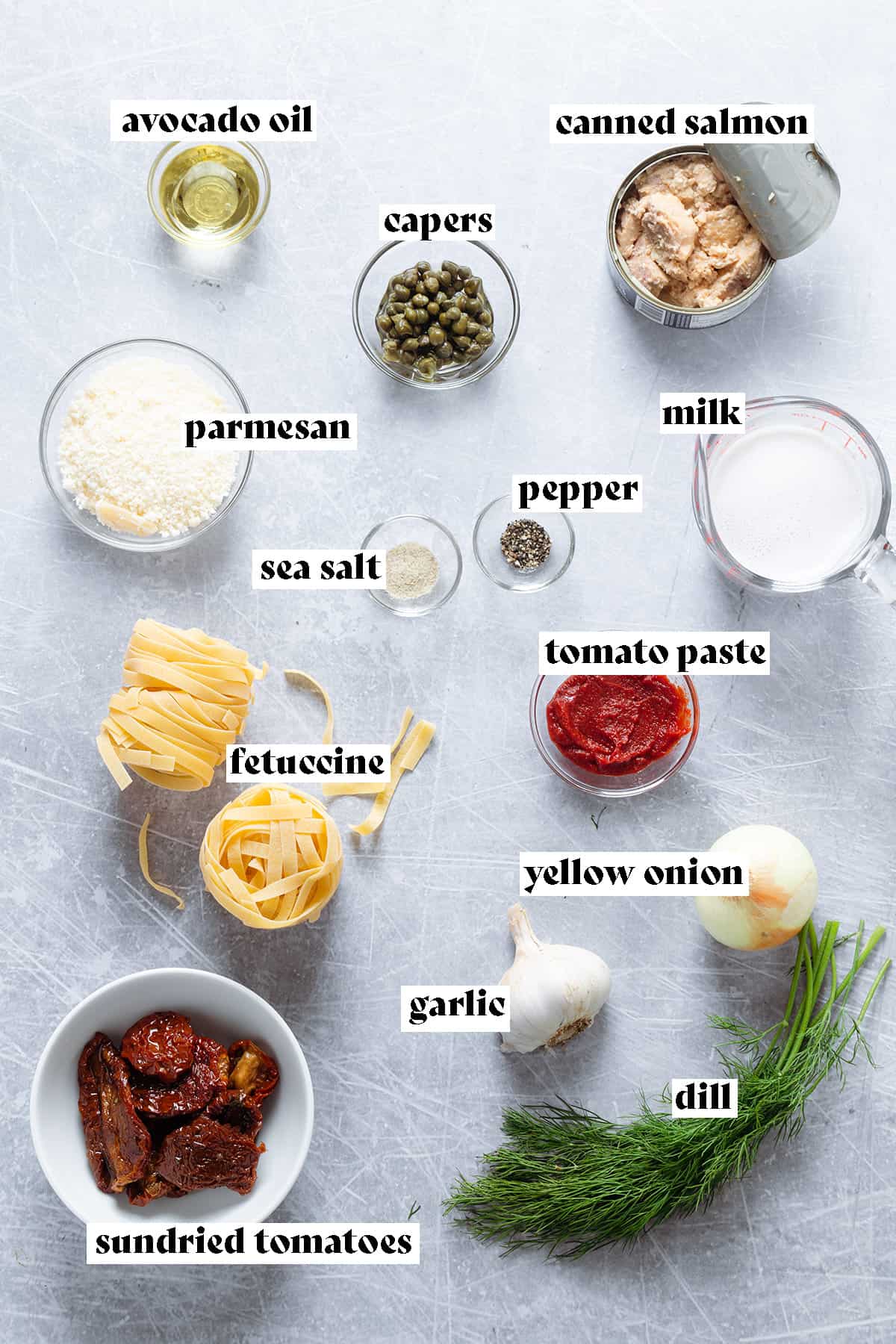 Ingredients for salmon pasta like fetuccine, sundried tomatoes, salmon, capers, and milk all laid out on a grey background.