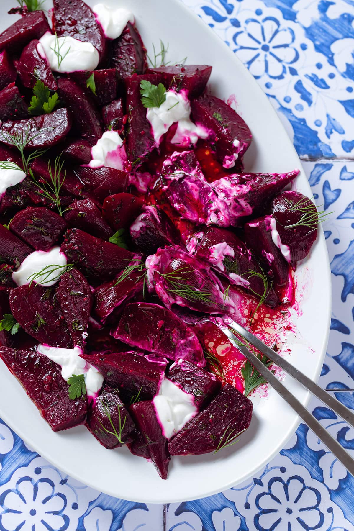 Beet salad with dollops of Greek yogurt and fresh herbs on a white serving platter with two serving spoons.