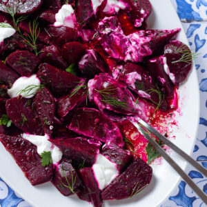 Beet salad with dollops of Greek yogurt partially mixed, and fresh herbs on a white serving platter with two serving spoons.