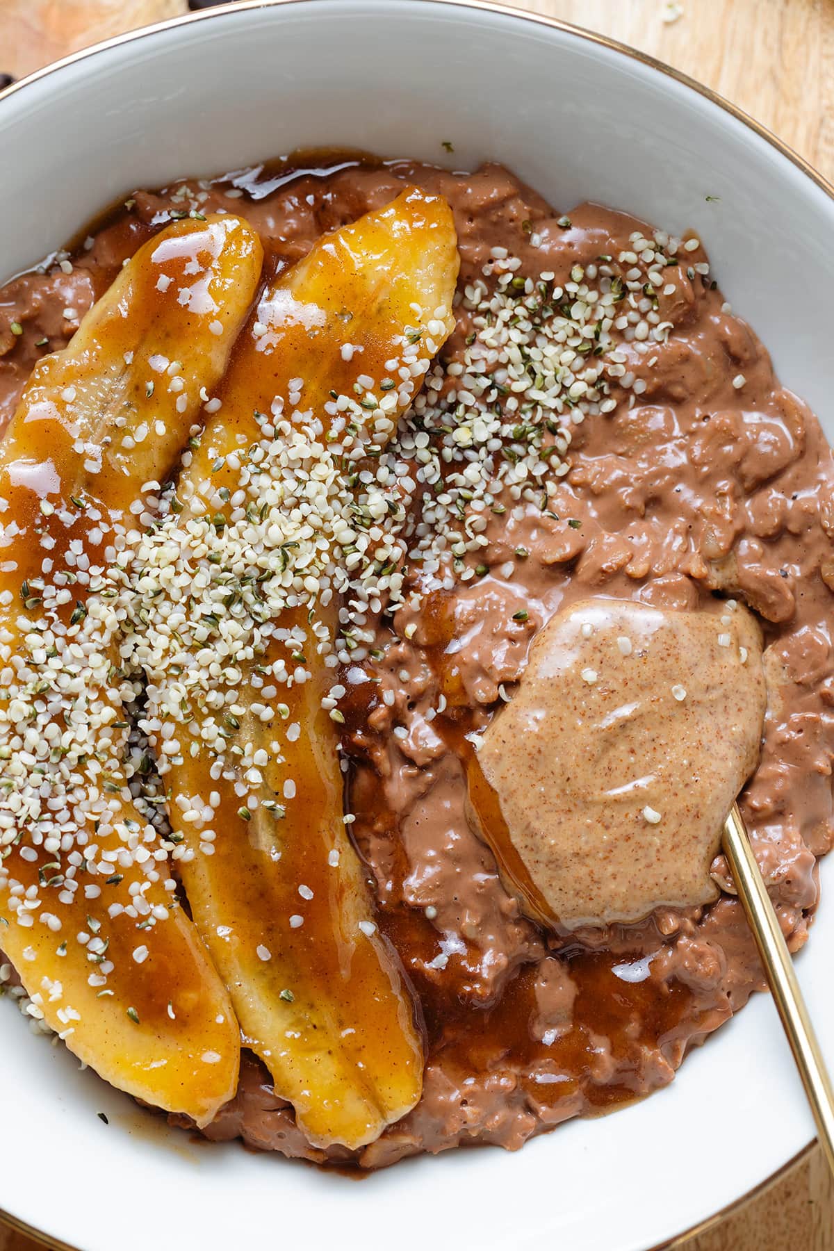 Chocolate coffee oatmeal in a white bowl topped with caramelized banana, almond butter, and hemp seeds with a gold spoon inserted on the right.