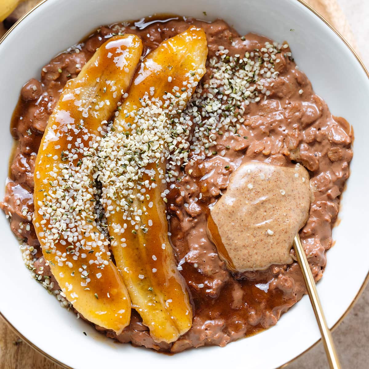 Chocolate coffee oatmeal in a white bowl topped with caramelized banana, almond butter, and hemp seeds with a gold spoon inserted on the right.