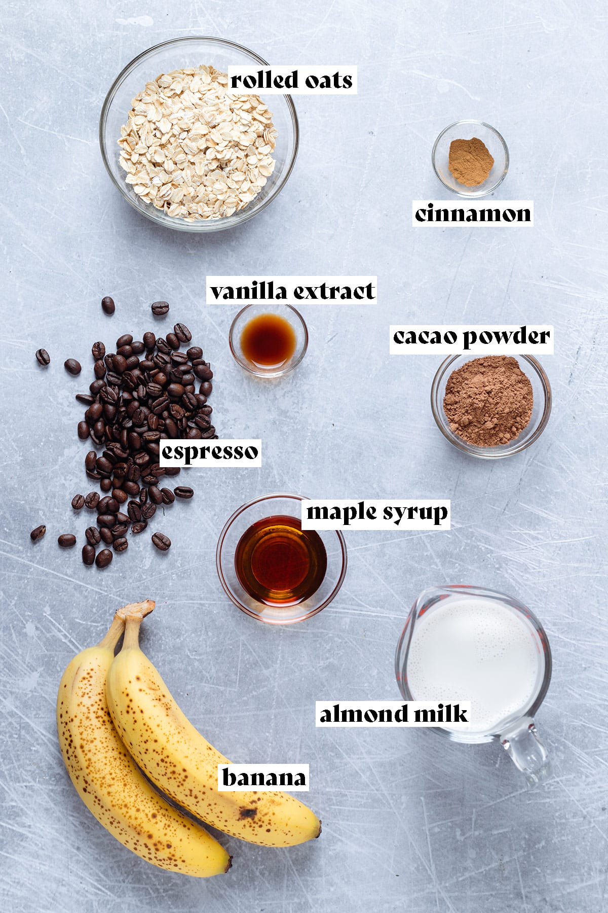 Ingredients for oatmeal like oats, bananas, maple syrup, cacao, milk, and other ingredients laid out on a grey background.