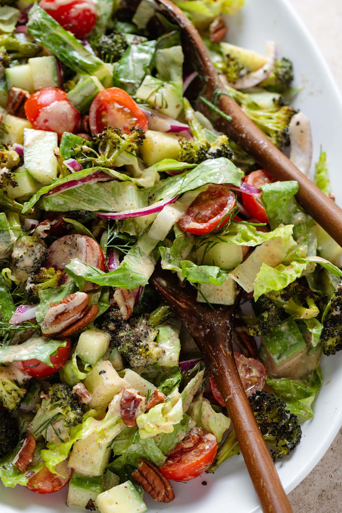 Chopped salad with roasted broccoli, romaine, cherry tomatoes, other vegetables, and creamy dressing on a white serving platter with two wooden spoons on the right.