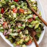 Chopped salad with roasted broccoli, romaine, cherry tomatoes, and other vegetables on a large white serving platter with two wooden spoons on the right.