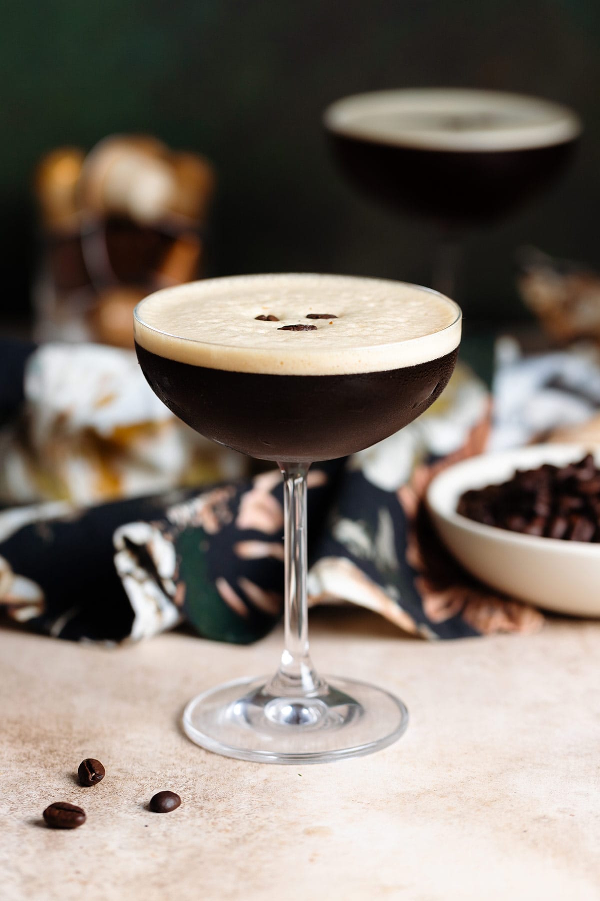 An espresso martini in a coupe glass with foam on top garnished with three coffee beans, another martini in the background, on a beige and dark green background.