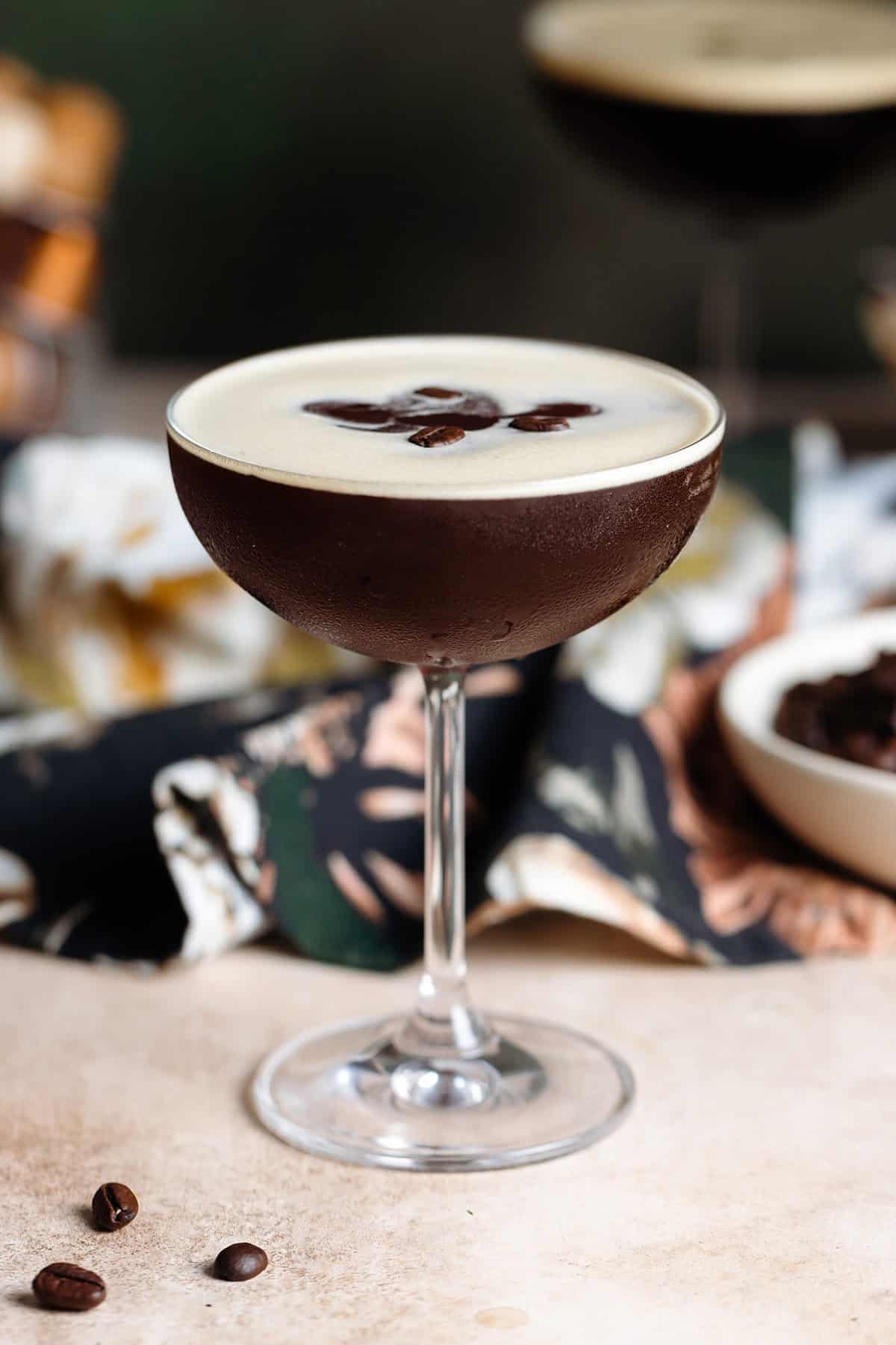 An espresso martini in a coupe glass with foam on top garnished with three coffee beans on a beige and dark green background.