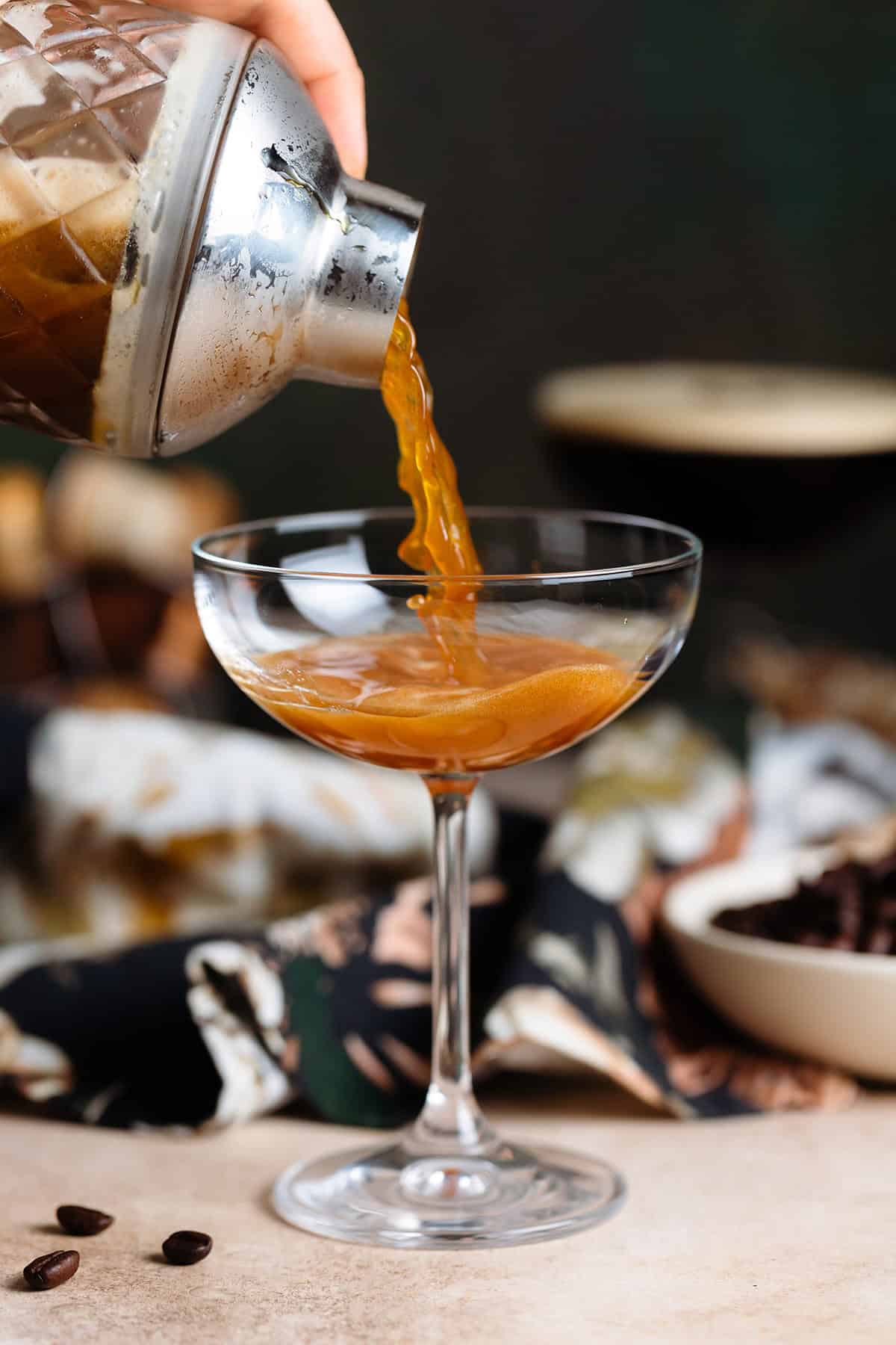 A tequila martini espresso being strained into a coupe glass from a cocktail shaker on a beige and dark green background.