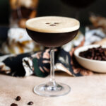 An espresso martini in a coupe glass with foam on top garnished with three coffee beans, another martini in the background, on a beige and dark green background.