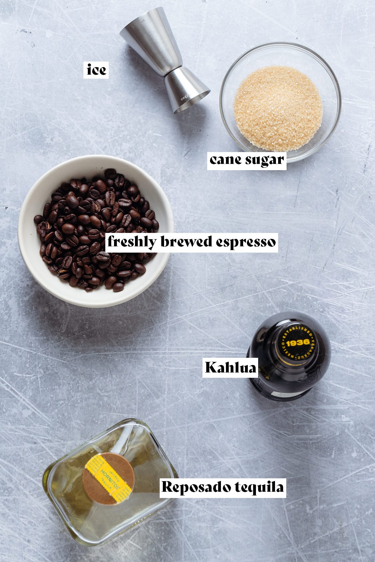 A bottle of tequila, Kahlua, cane sugar, and espresso beans laid out on a grey background.