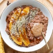 Chocolate coffee oatmeal in a white bowl topped with caramelized banana, almond butter, and hemp seeds with a gold spoon inserted on the right.