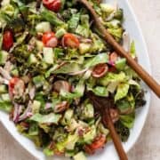 Chopped salad with roasted broccoli, romaine, cherry tomatoes, and other vegetables on a large white serving platter with two wooden spoons on the right.