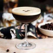 An espresso martini in a coupe glass with foam on top garnished with three coffee beans, another martini in the background, on a beige and dark green background.