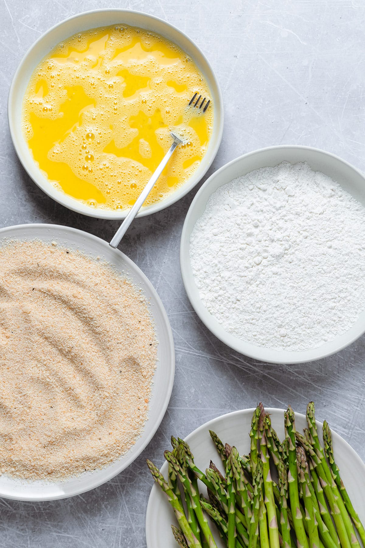 Three plates with eggs, flour, and breadcrumbs ready to batter asparagus.