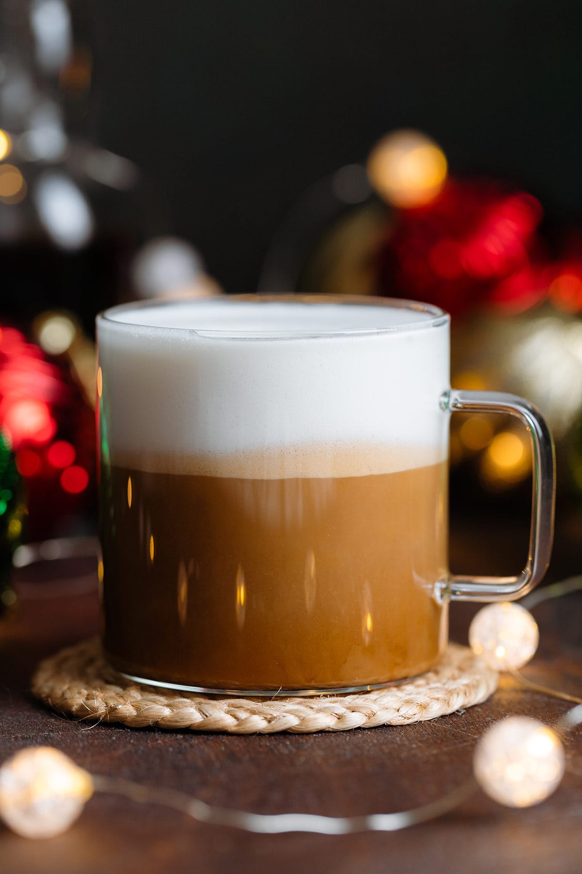 A chestnut praline latte in a glass mug on a dark wooden background with Christmas ornaments behind it.