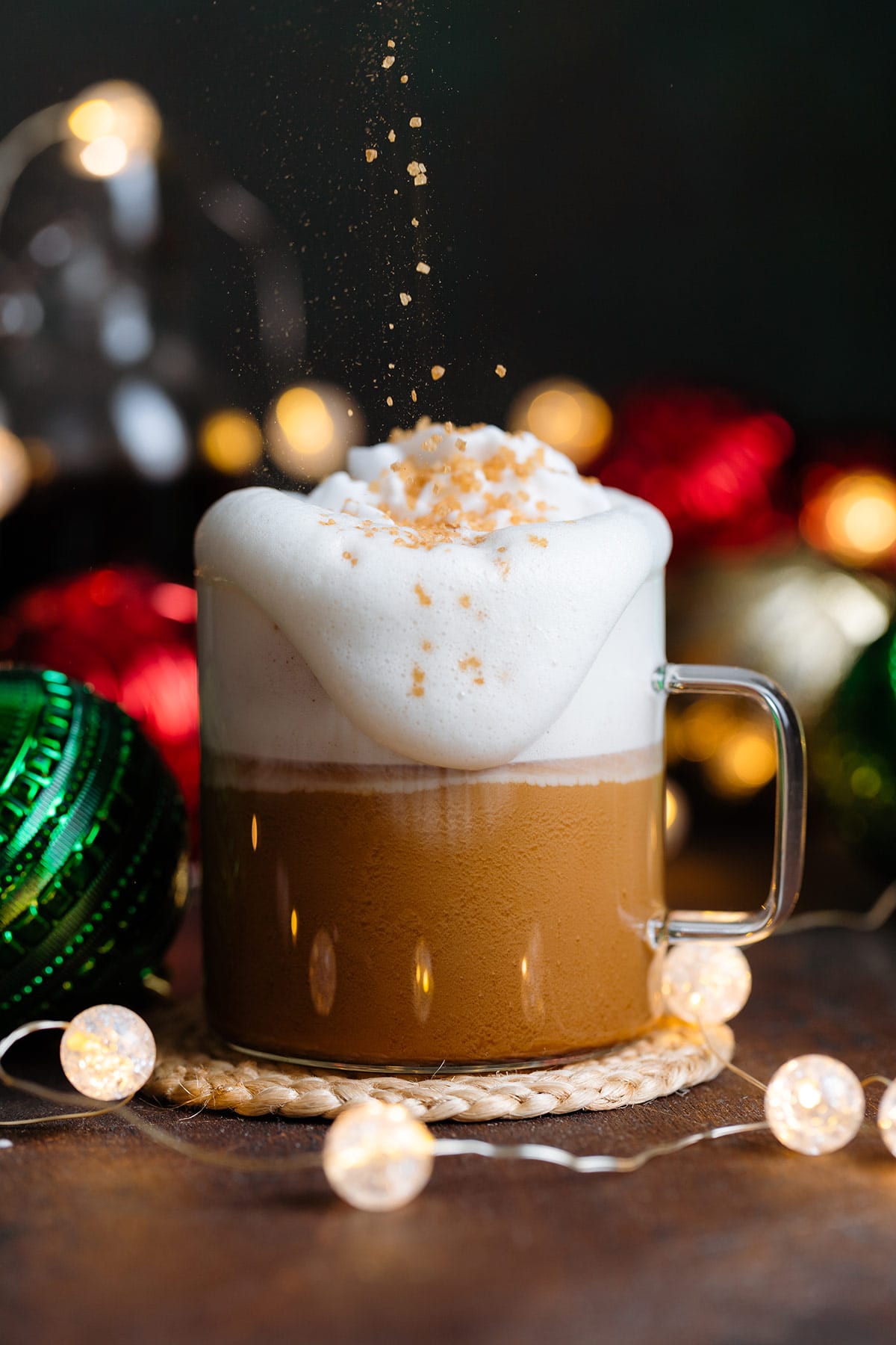A cafe latte overflowing in a glass mug with whipped cream and coarse sugar on top on a dark wooden background with Christmas ornaments around.