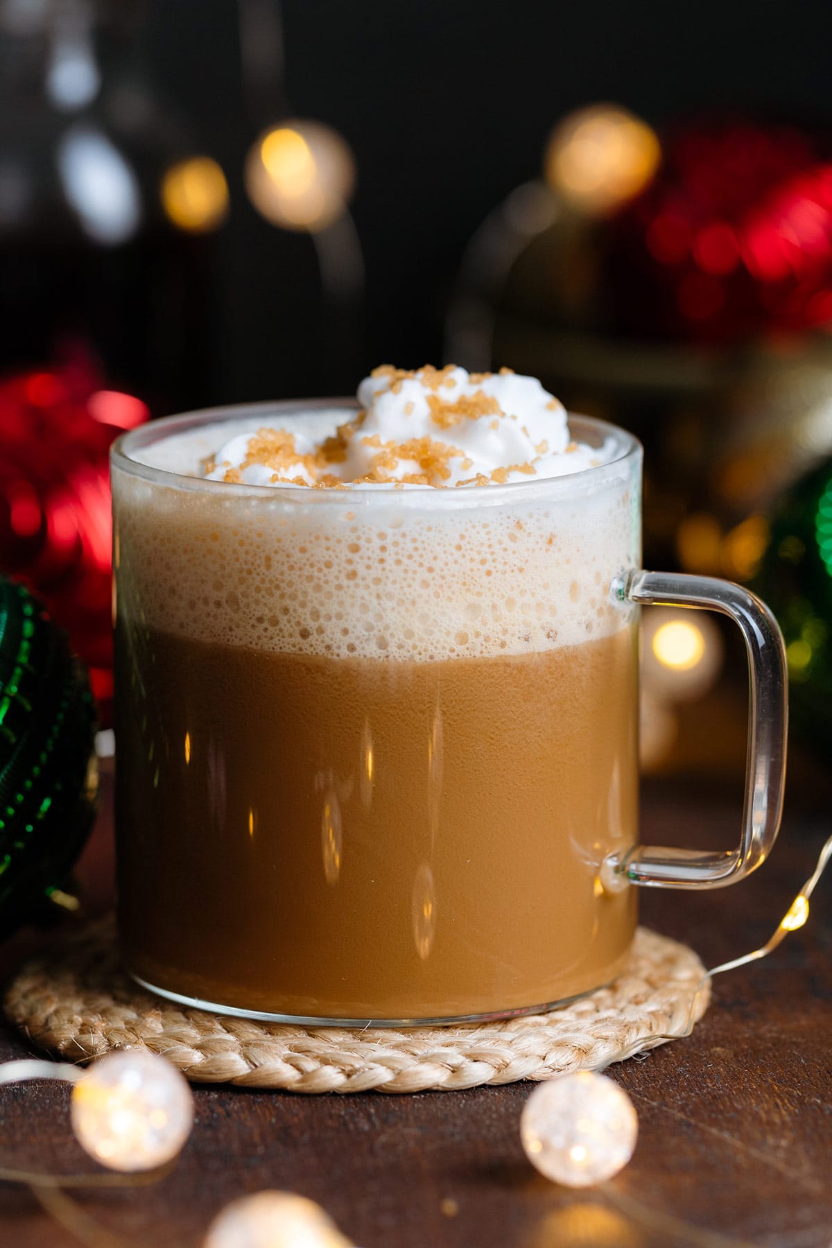 A cafe latte in a glass mug with whipped cream and coarse sugar on top on a dark wooden background with Christmas ornaments around.