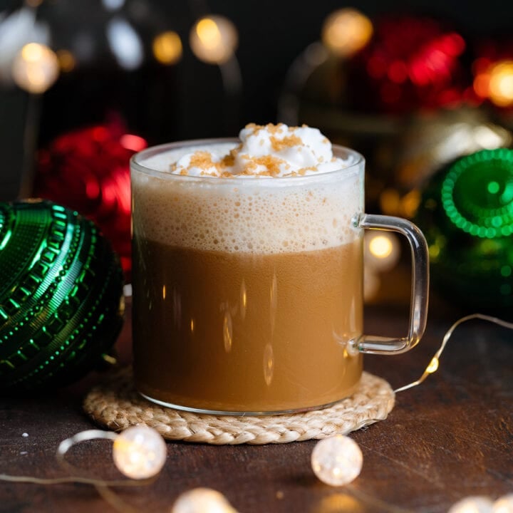 A cafe latte in a glass mug with whipped cream and coarse sugar on top on a dark wooden background with Christmas ornaments around.