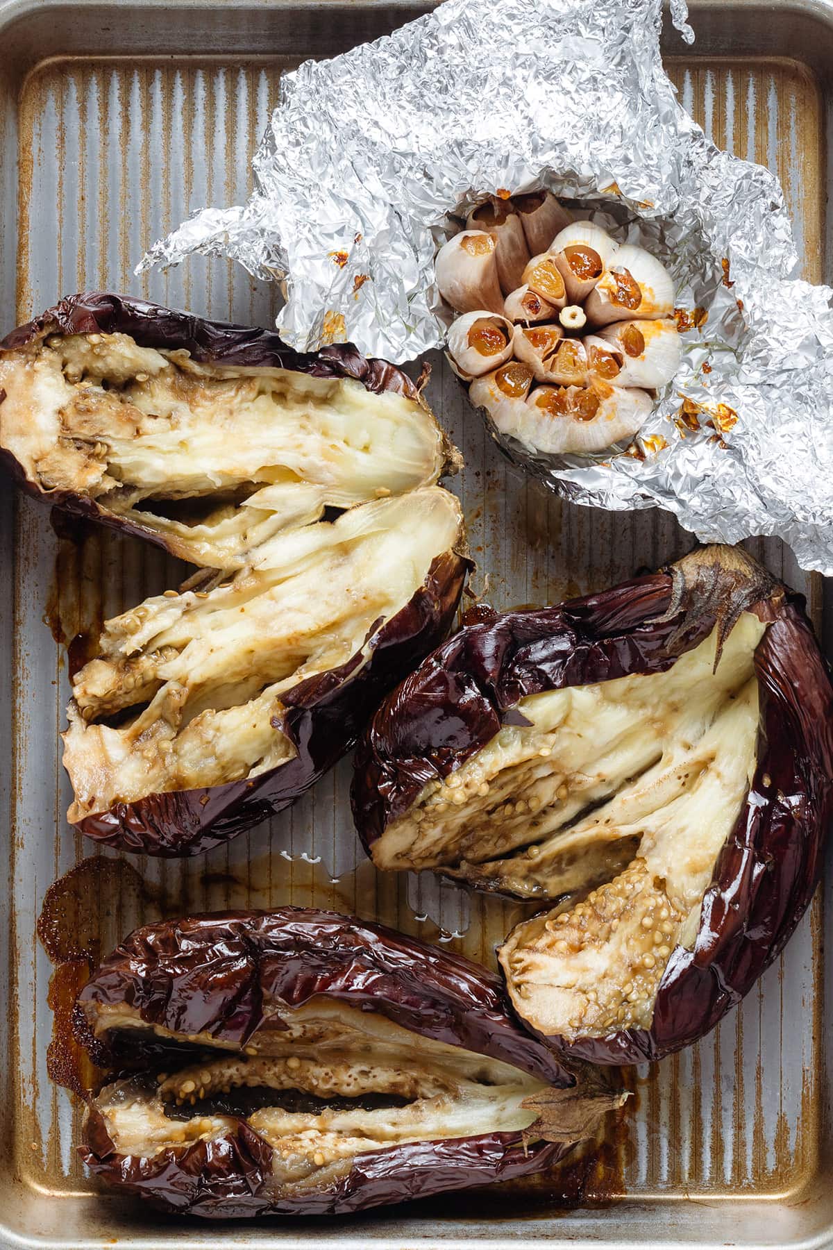 Roasted whole eggplants sliced in half on a baking sheet with roasted garlic in foil.