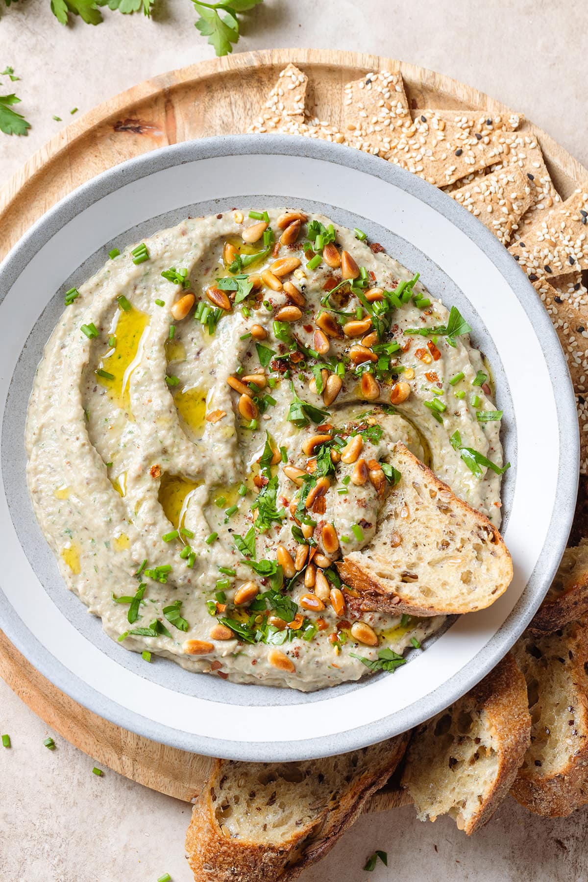 Creamy eggplant dip in a grey low bowl with a white edge, topped with toasted pine nuts and fresh herbs, with a slice of a toasted baguette dipped into the dip.