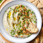 Creamy eggplant dip in a grey low bowl with a white edge, topped with toasted pine nuts and fresh herbs served with a toasted baguette on the side.