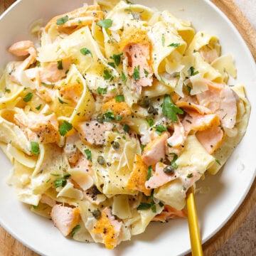 Pappardelle with roasted salmon, creamy white sauce, black pepper, and fresh herbs on a white plate.