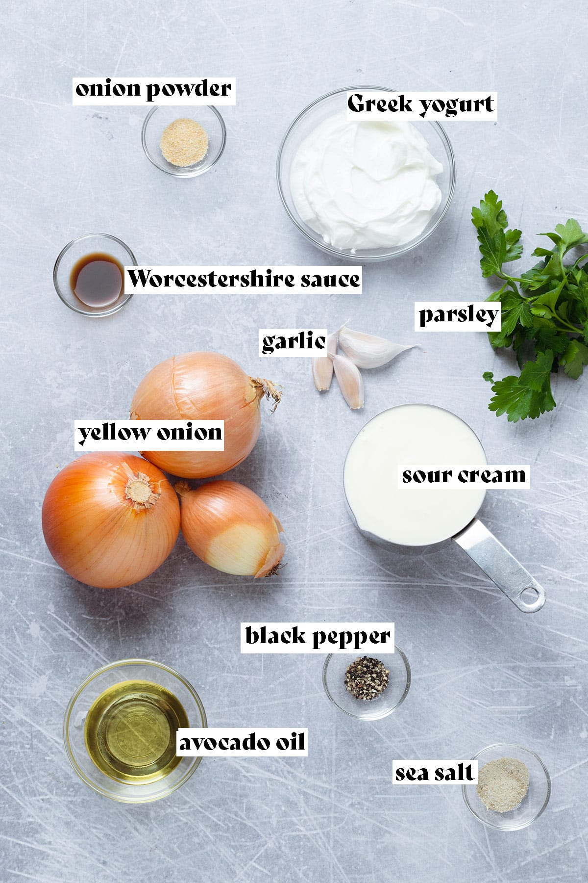 Onions, garlic, Greek yogurt, sour cream, fresh parsley, and other ingredients laid out on a grey background.