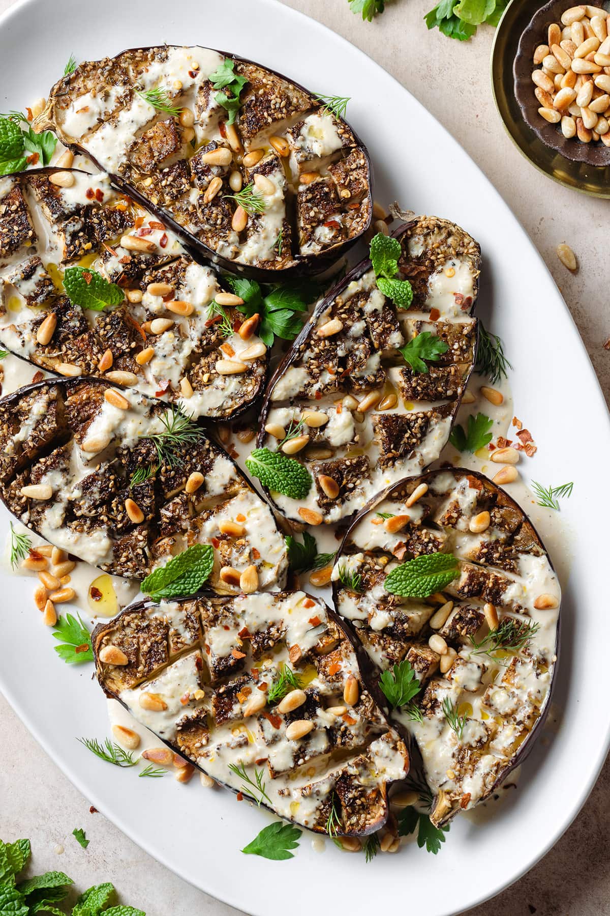 Halved roasted eggplant with spices, tahini sauce, herbs, and pine nuts on a large white serving plate.