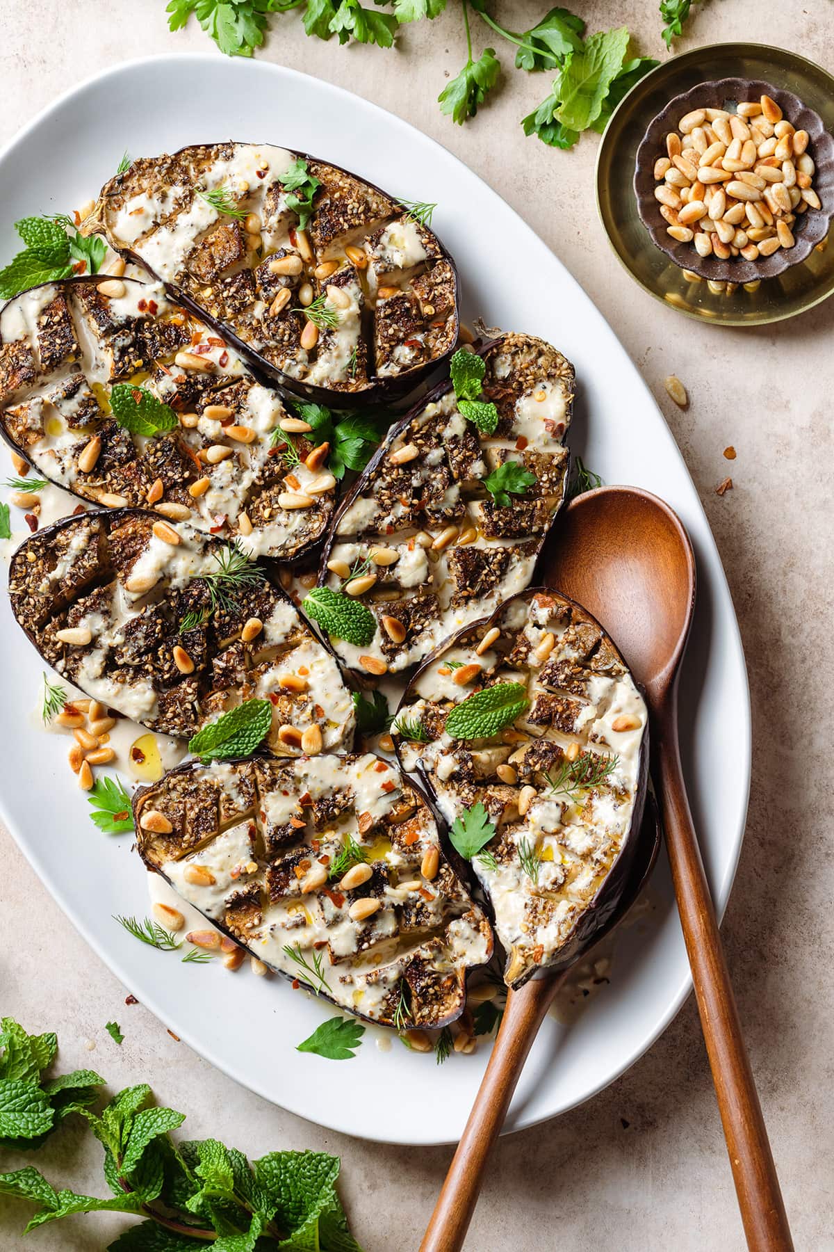 Halved roasted eggplant with spices, tahini sauce, herbs, and pine nuts on a large white plate with wooden serving spoons on the right.