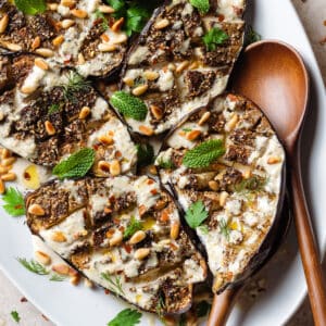 Halved roasted eggplant with spices, tahini sauce, herbs, and pine nuts on a large white plate with wooden serving spoons on the right.