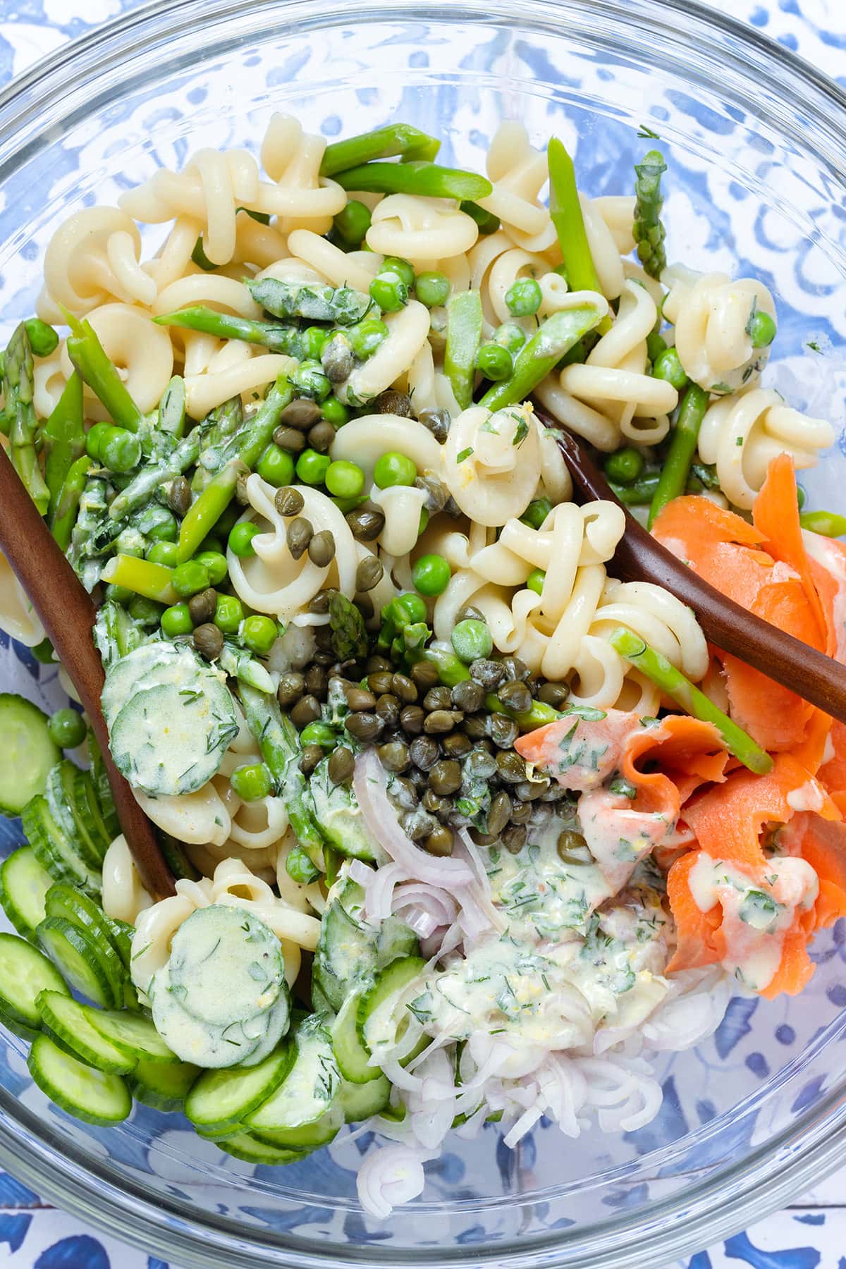 Pasta salad with smoked salmon, asparagus, peas, cucumber, shallots, capers, and creamy dressing in a large glass bowl.