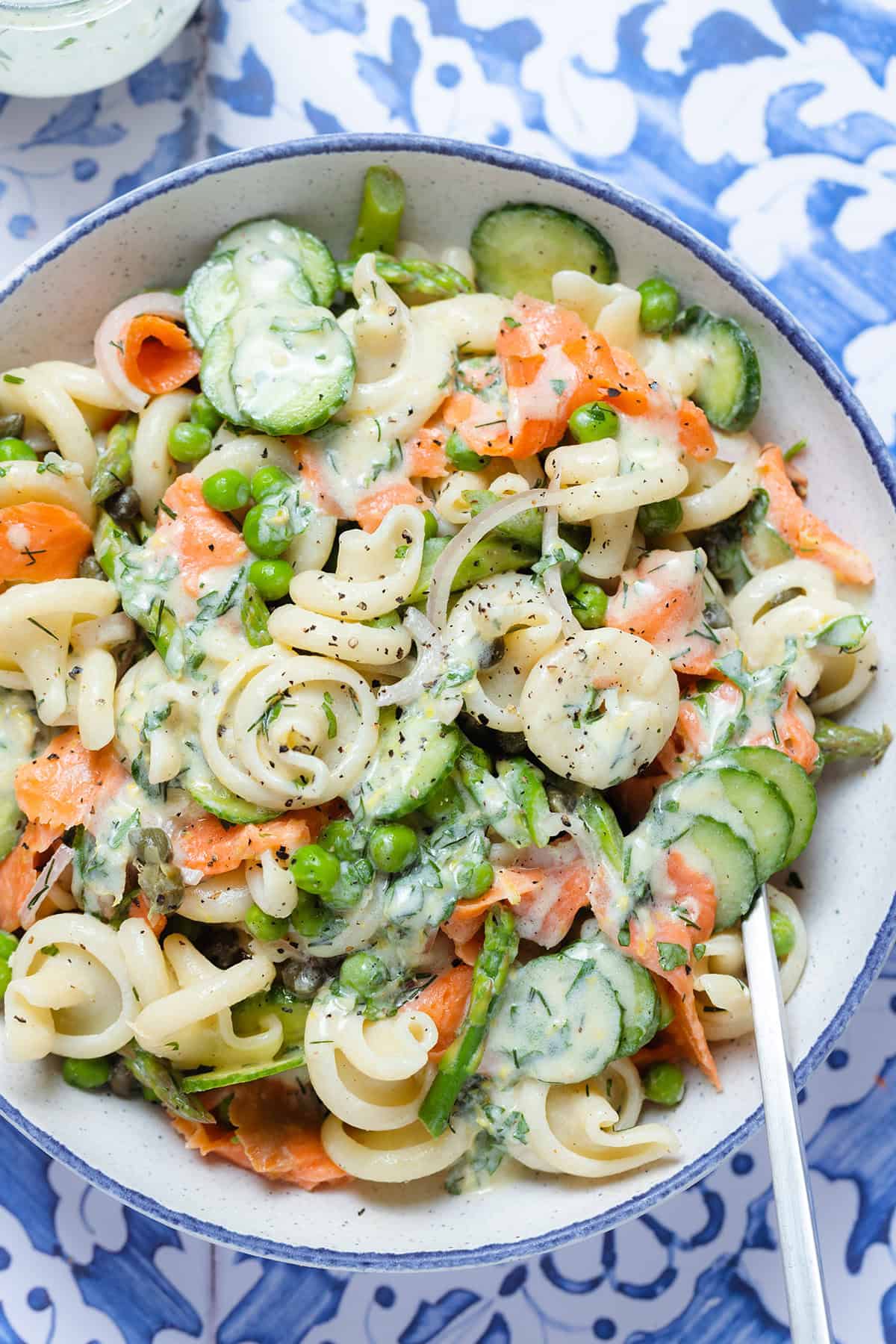 Creamy pasta salad with smoked salmon, peas, and asparagus in a white bowl on a blue tile background.