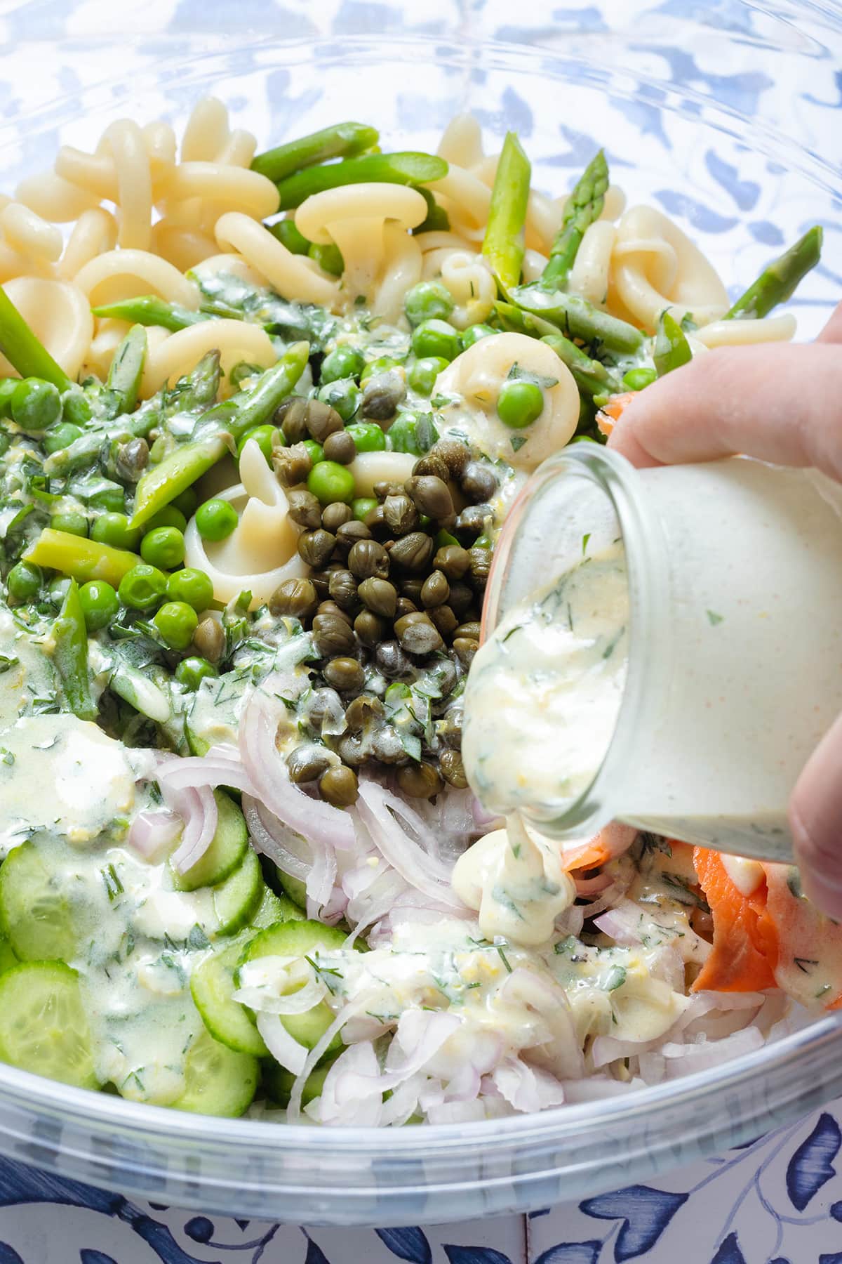 A pasta salad with smoked salmon in a large glass bowl with creamy herb dressing being poured over it.
