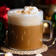 A cafe latte in a glass mug with whipped cream and coarse sugar on top on a dark wooden background with Christmas ornaments around.