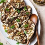 Halved roasted eggplant with spices, tahini sauce, herbs, and pine nuts on a large white plate with wooden serving spoons on the right.