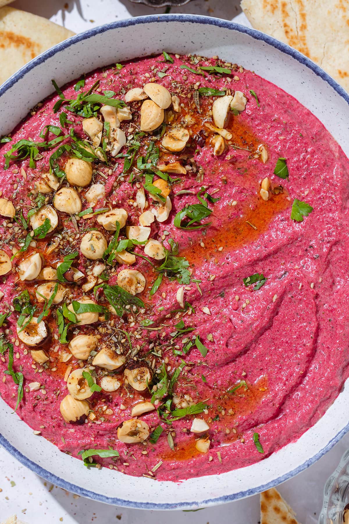 Bright pink beet dip in a white bowl with a blue rim garnished with hazelnuts, parsley, and olive oil.