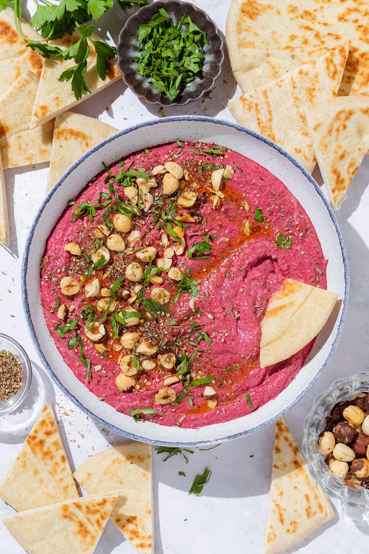 Bright pink beet dip in a white bowl with a blue rim garnished with hazelnuts, parsley, and olive oil with a naan triangle dipped into the dip.