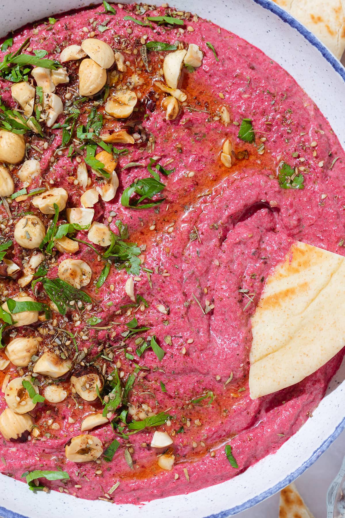 Bright pink beet dip in a white bowl with a blue rim garnished with hazelnuts, parsley, and olive oil with a naan triangle dipped into the dip.