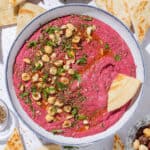 Bright pink beet dip in a white bowl with a blue rim garnished with hazelnuts, parsley, and olive oil with a naan triangle dipped into the dip.