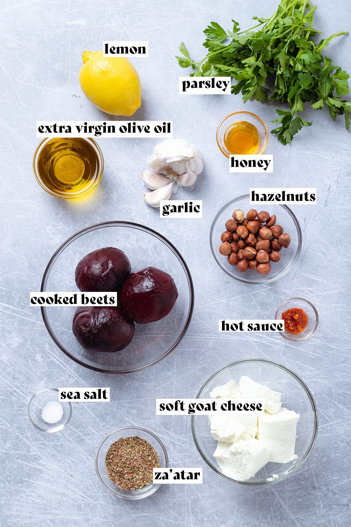 Ingredients like cooked beets, goat cheese, lemon, parsley, garlic, and hazelnuts laid out on a grey background.