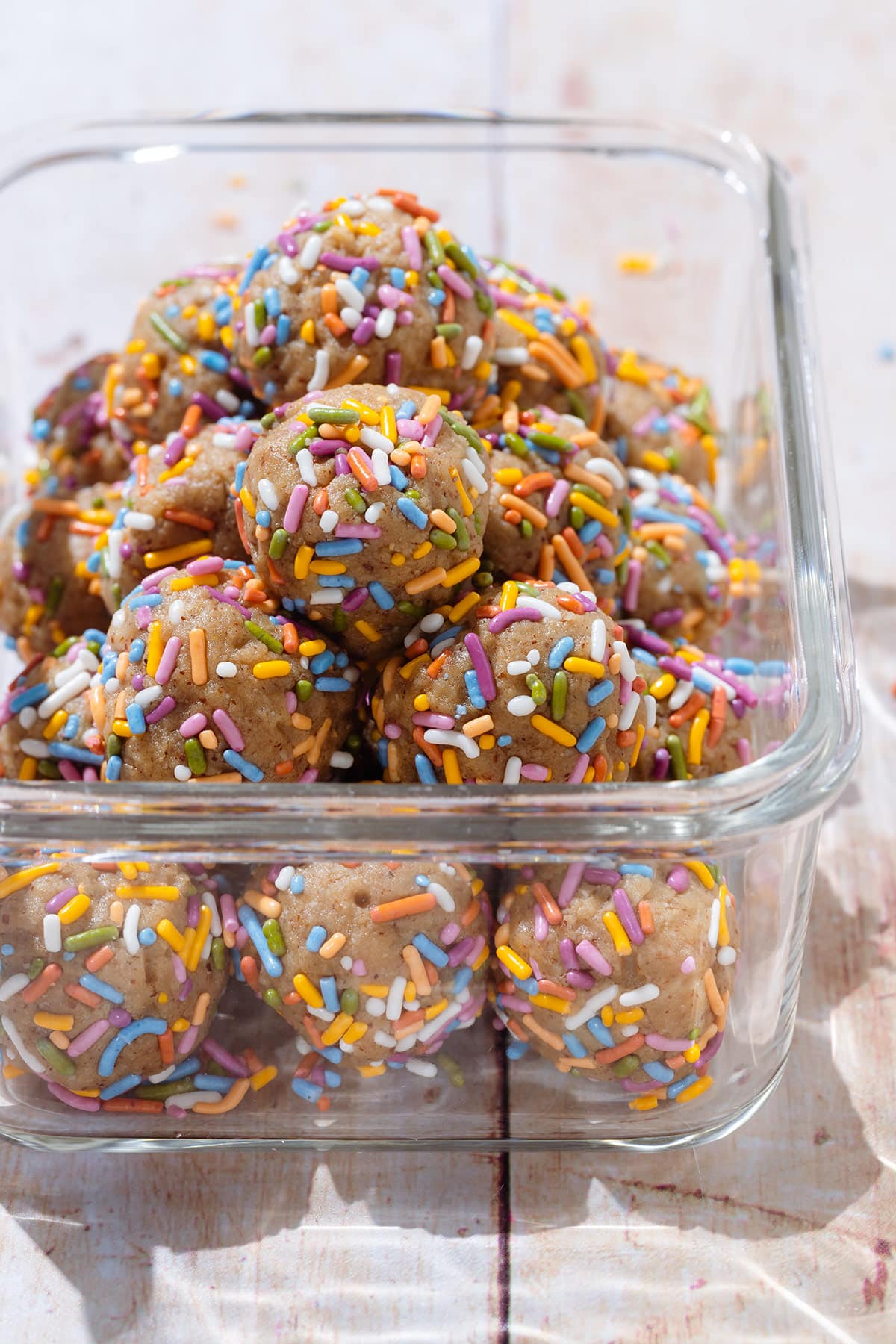 Protein bites covered with multi-colored sprinkles in a glass container on a light wooden background.