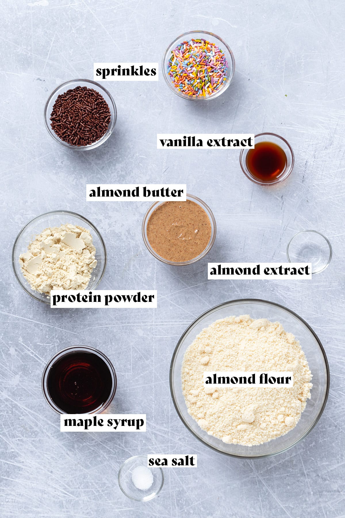 Ingredients in small bowls for birthday cake protein balls laid out on a light grey background.