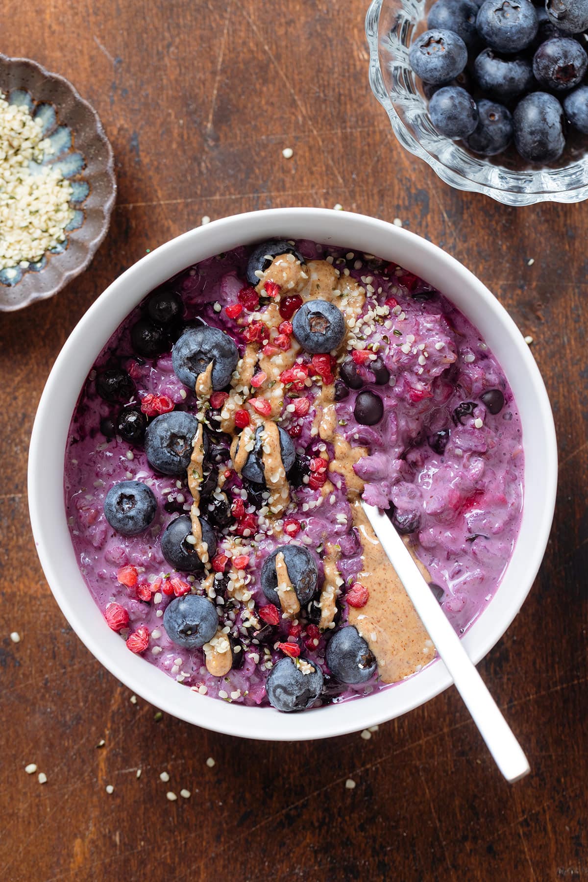 Bright purple blueberry oatmeal garnished with blueberries, nut butter, and hemp seeds on a dark wooden background.
