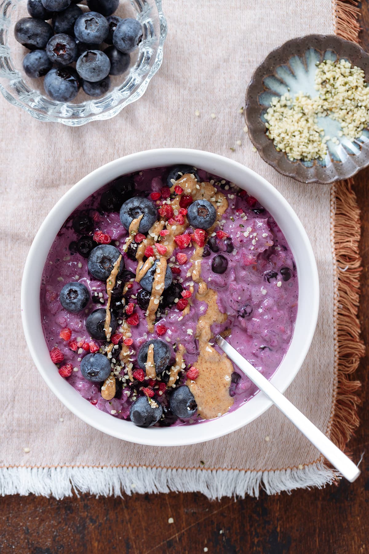 Bright purple blueberry oatmeal in a white bowl garnished with blueberries, nut butter, and hemp seeds on a dark wooden background and a beige tablecloth.