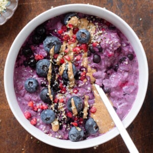 Bright purple blueberry oatmeal in a white bowl garnished with blueberries, nut butter, and hemp seeds on a dark wooden background.