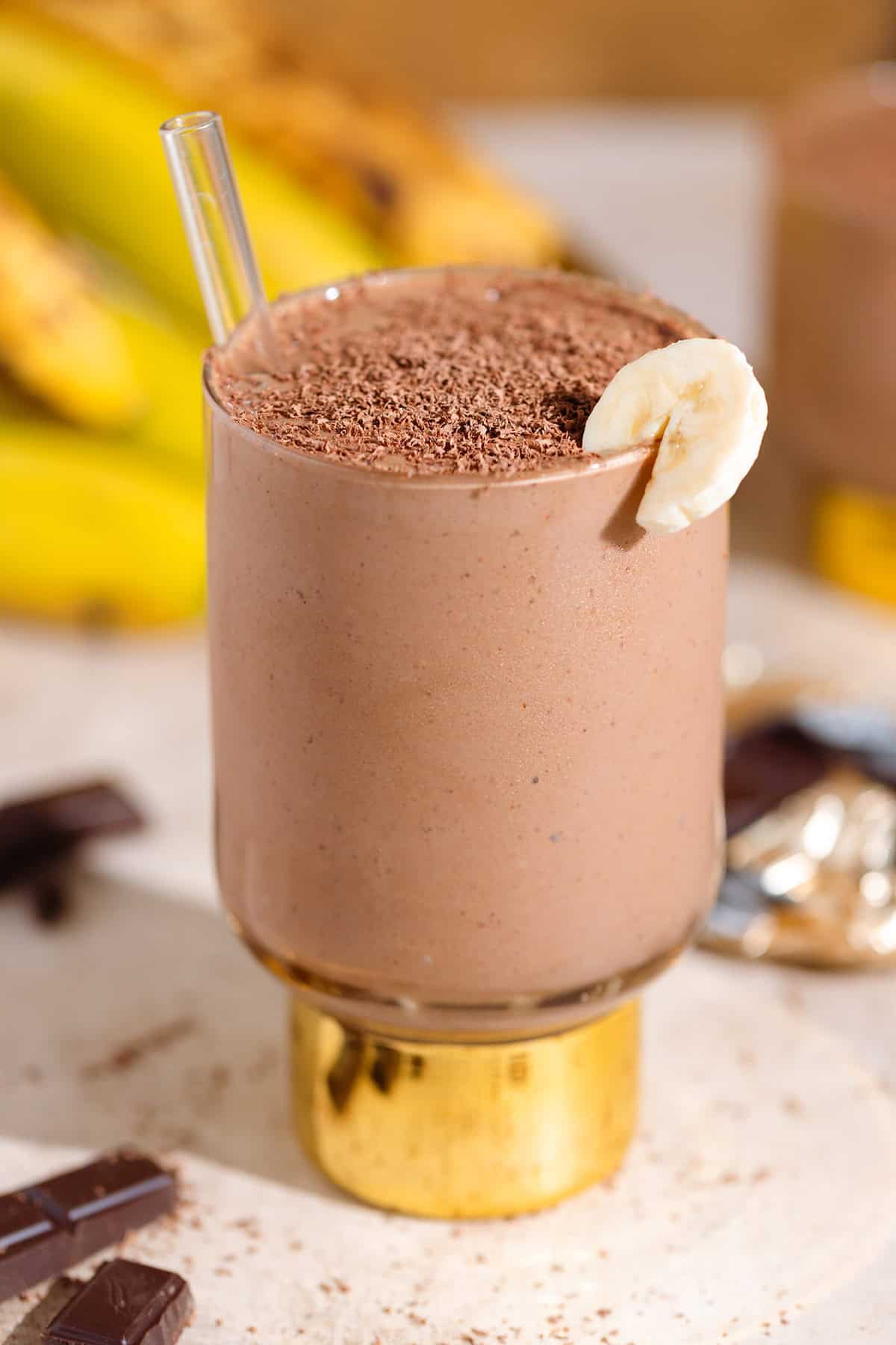 Chocolate smoothie in a tall glass with a shiny gold thick stem garnished with a slice of banana and shaved chocolate.