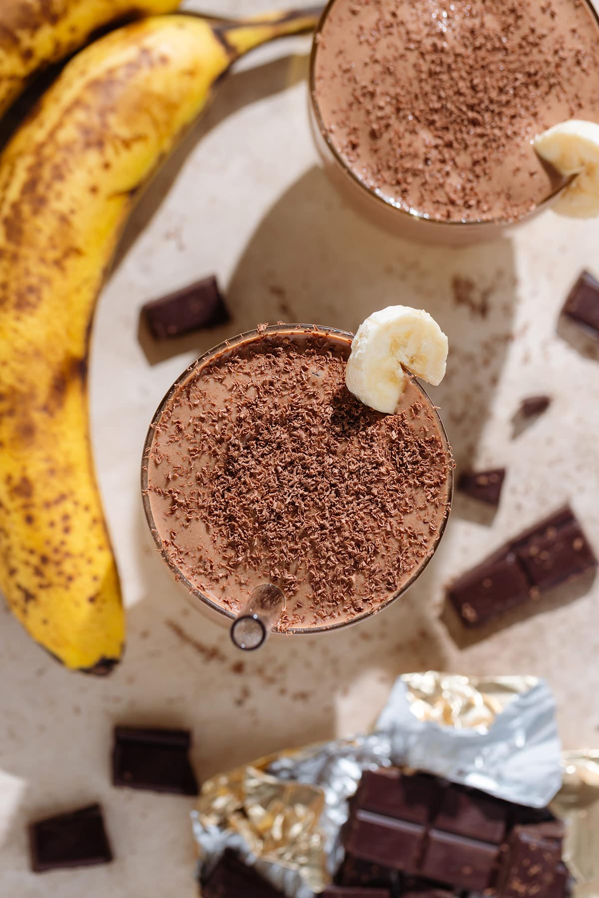 Chocolate smoothie in a tall glass garnished with a slice of banana and shaved chocolate.