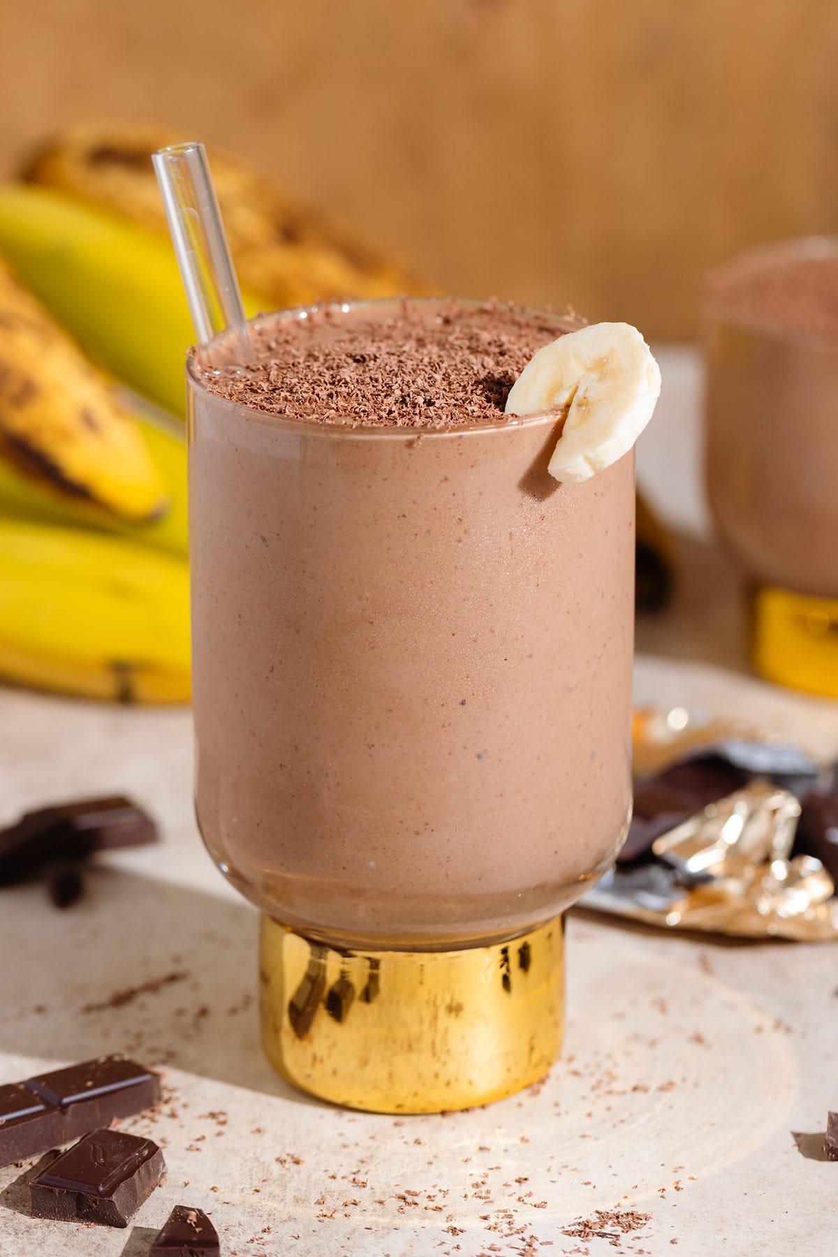 Chocolate smoothie in a tall glass with a shiny gold thick stem garnished with a slice of banana and shaved chocolate.