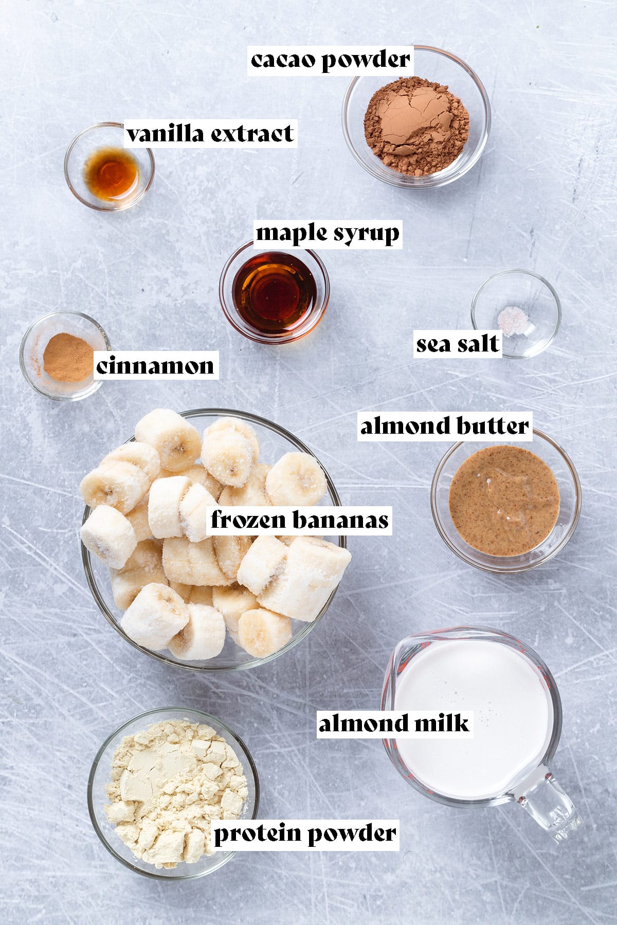 Ingredients like frozen bananas, maple syrup, almond butter, and cacao laid out on a grey background with text overlay.