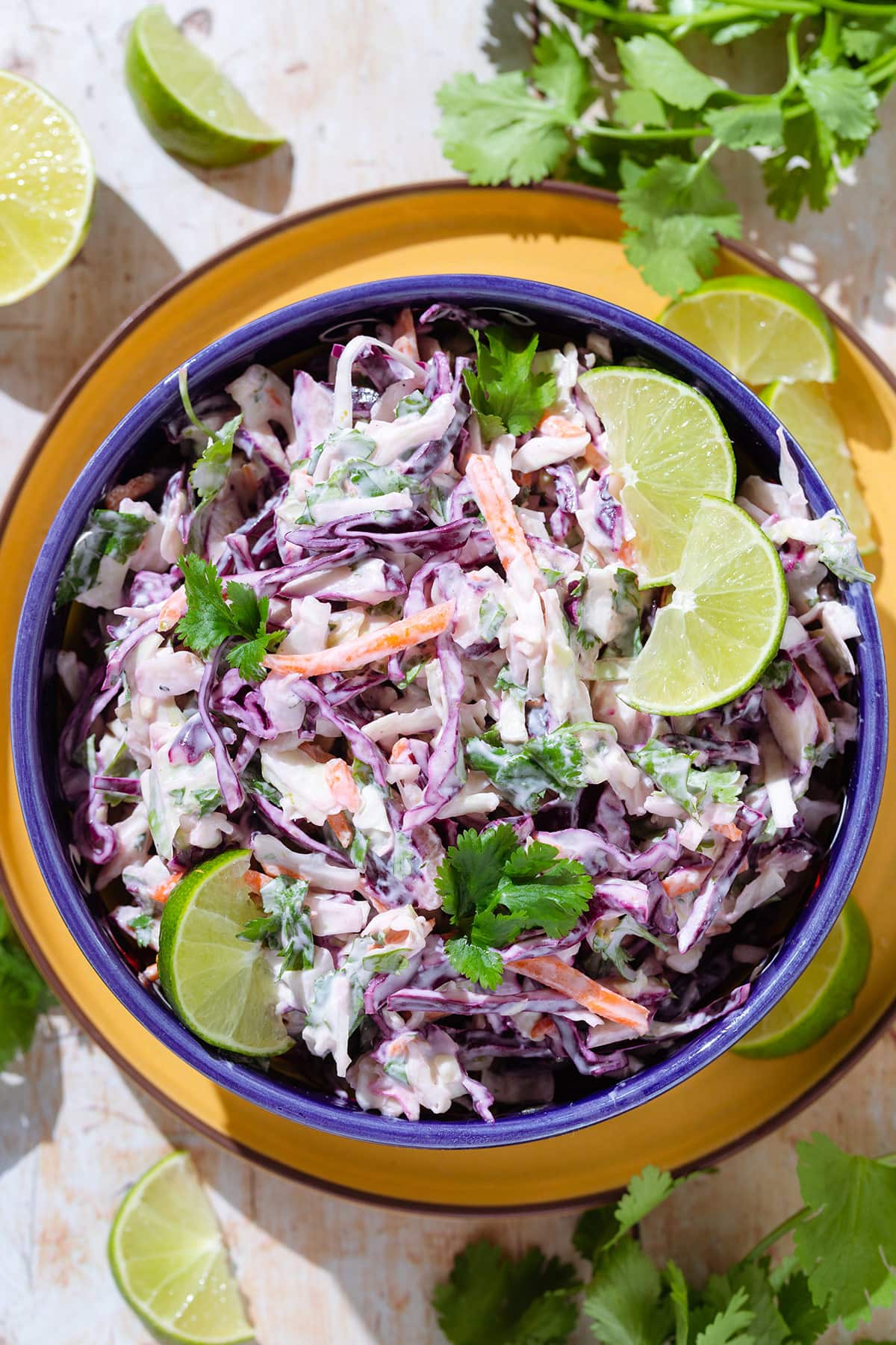 Colorful cilantro lime slaw with purple cabbage in a blue bowl garnished with lime slices on a yellow plate.
