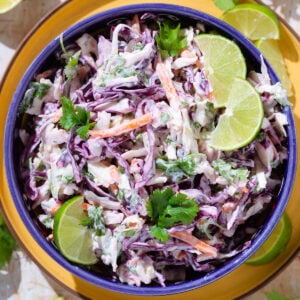 Colorful cilantro lime slaw with purple cabbage in a blue bowl garnished with lime slices on a yellow plate.